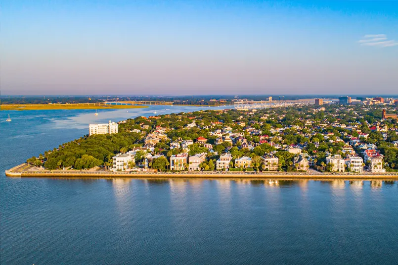Aerial view of a coastal city with historic homes, surrounded by water and greenery under a clear blue sky.
