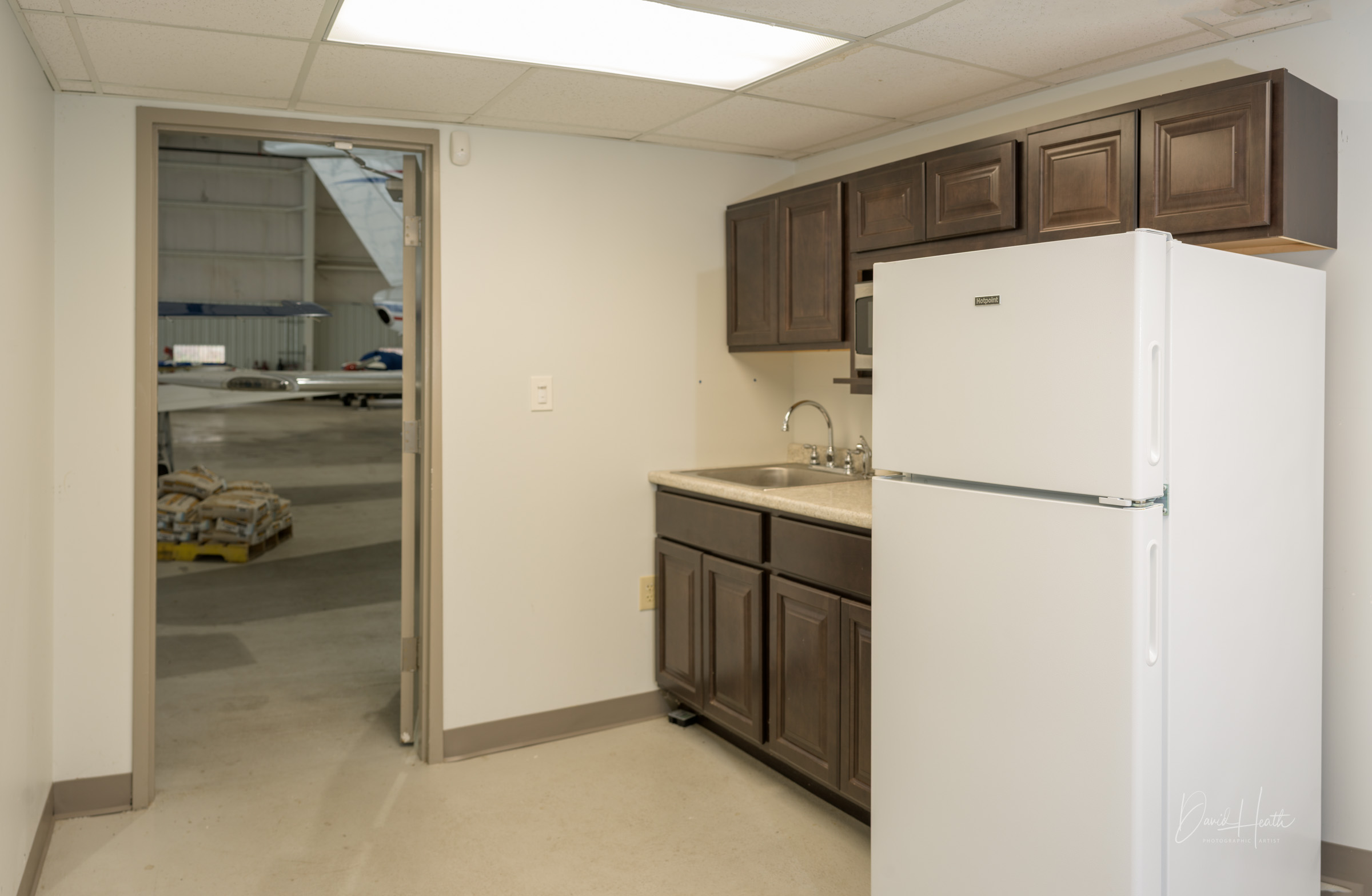 Small kitchenette with a fridge, sink, and dark cabinets in an industrial setting near an open door to an airplane hangar.