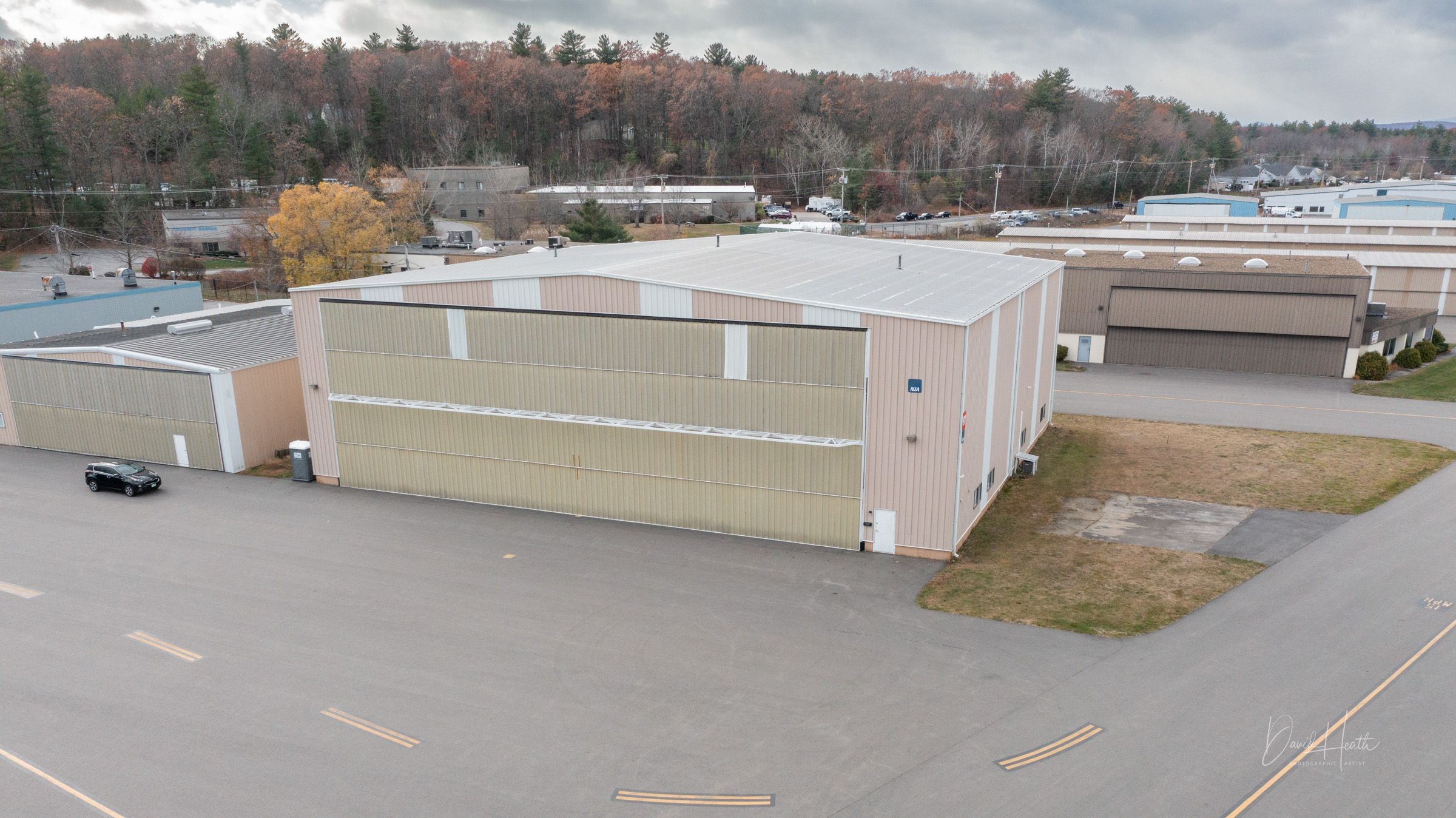 Aerial view of a large industrial warehouse near a forest, featuring a parked car and overcast skies.